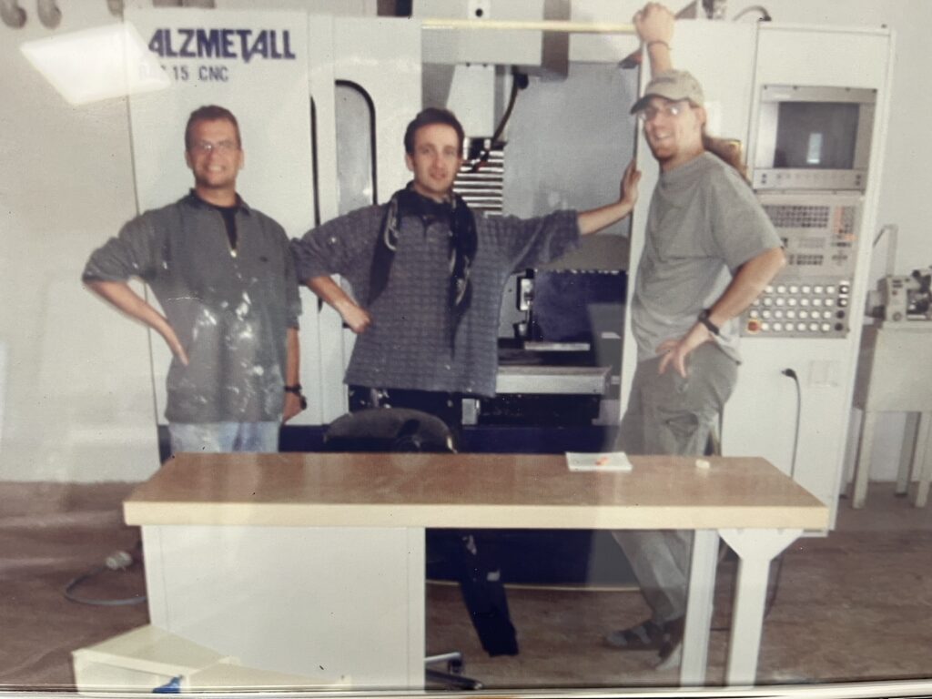 Bei der Gründung 2004 posieren drei Mitarbeiter vor einer CNC-Maschine in der Werkstatt von HFH Präzisionsmechanik GmbH.