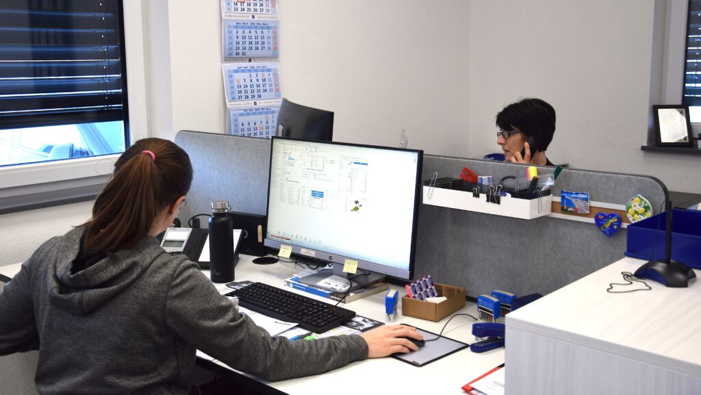 Im Büro der Dritten Halle von HFH Präzisionsmechanik GmbH arbeitet eine Mitarbeiterin am Computer, während im Hintergrund ein Kollege telefoniert.