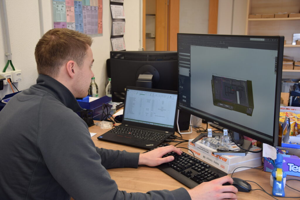 Ein Mitarbeiter arbeitet an der Konstruktion und CAD/CAM-Planung mit SolidWorks an zwei Monitoren und einem Laptop im Büro.
